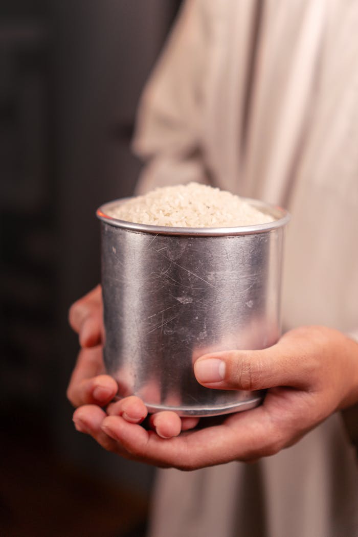 Close-up of hands holding a tin cup full of uncooked white rice, symbolizing abundance.